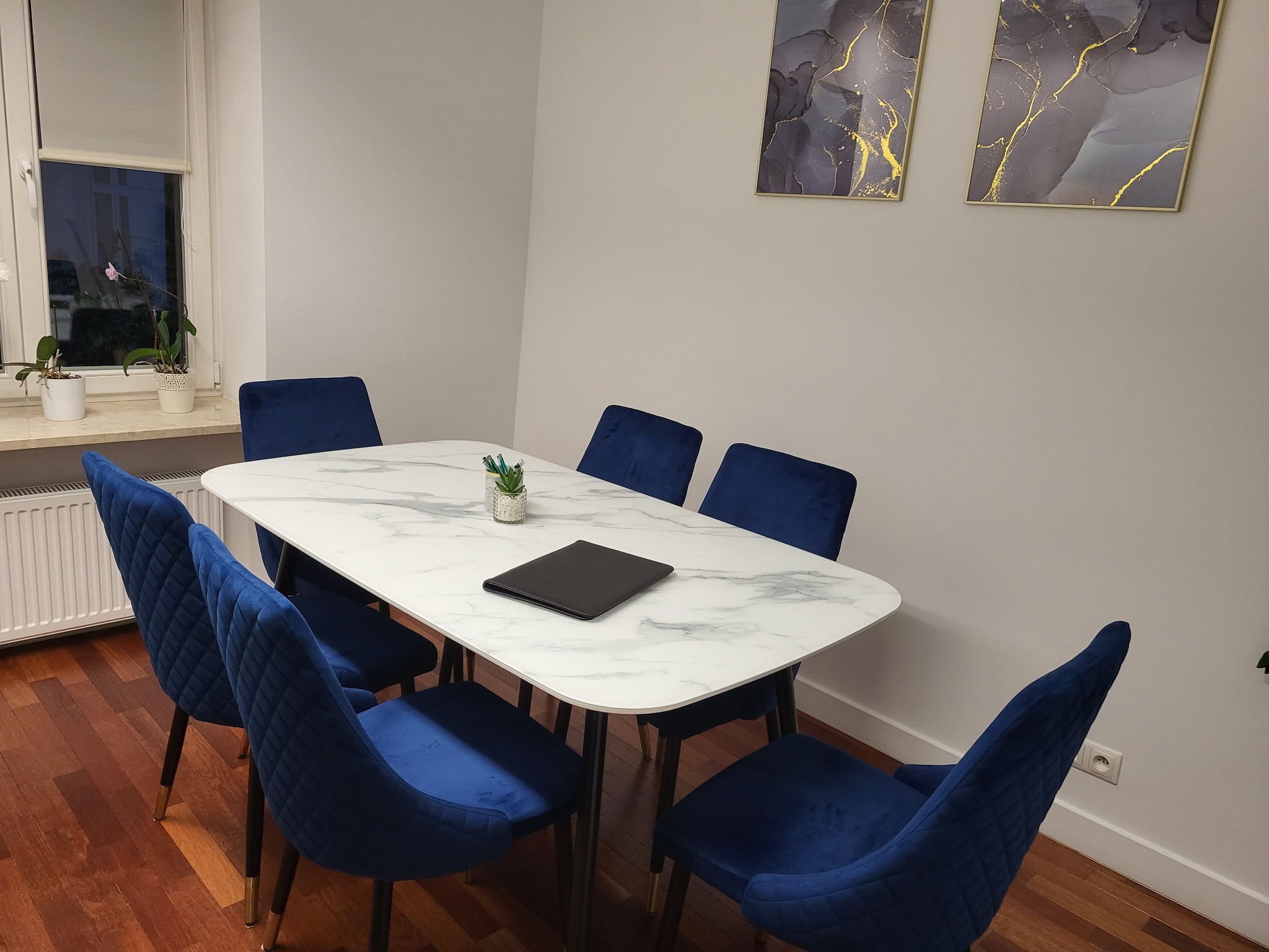 Sala konferencyjna z marmurowym stołem, sześcioma granatowymi krzesłami, czarnym notatnikiem i rośliną w ceramicznej doniczce. Na ścianie wiszą dwa abstrakcyjne obrazy w złotych ramkach.