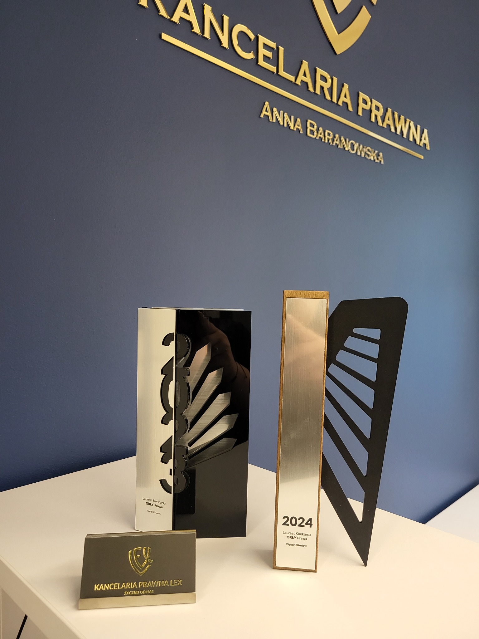 Nagrody i wizytówka Kancelarii Prawnej Anny Baranowskiej na białym stole przed niebieską ścianą z napisem Kancelaria Prawna.