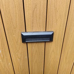 TM SYSTEM - Zamknięty, czarny wideodomofon na brązowych, pionowych panelach drewnopodobnych. Nowoczesny design, zbliżenie na detal. Minimalistyczny styl.