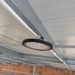 TM SYSTEM - Okrągła lampa LED zamontowana pod sufitem z blachy trapezowej w surowym wnętrzu. Widoczne metalowe profile konstrukcyjne i instalacja elektryczna.