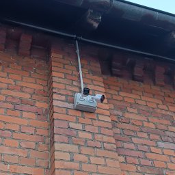 TM SYSTEM - Kamera monitoringu zamontowana na ceglanej ścianie budynku, z widocznymi przewodami w peszlu i puszką instalacyjną. Ujęcie z dołu, widoczny fragment rynny.