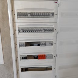 TM SYSTEM - Otwarta, biała skrzynka elektryczna z zainstalowanymi bezpiecznikami Eaton i ETI, widoczna na tle szarej ściany. Układ bezpieczników wewnątrz, widoczne przewody.