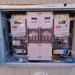 TM SYSTEM - Otwarta skrzynka z dwoma licznikami energii elektrycznej i zabezpieczeniami, widoczne przewody, kostki elektryczne i elementy montażowe. Instalacja w szarej obudowie na szarej ścianie.