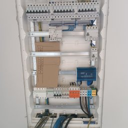 TM SYSTEM - Rozdzielnica elektryczna wnękowa, z zainstalowanymi bezpiecznikami, zabezpieczeniami przepięciowymi i okablowaniem. Widoczne brązowe kartki z opisem obwodów.