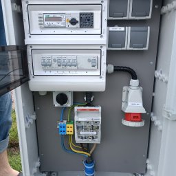 TM SYSTEM - Otwarta skrzynka elektryczna z bezpiecznikami, gniazdami (1-230V, 2-230V, -400V), licznikiem energii i okablowaniem, widoczne elementy instalacji elektrycznej.