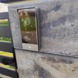 Nowoczesny wideodomofon EZVIZ zamontowany na kamiennym słupku bramy wjazdowej, widoczny z bliska. Słup z szaroburego kamienia, brama metalowa, w tle dom.