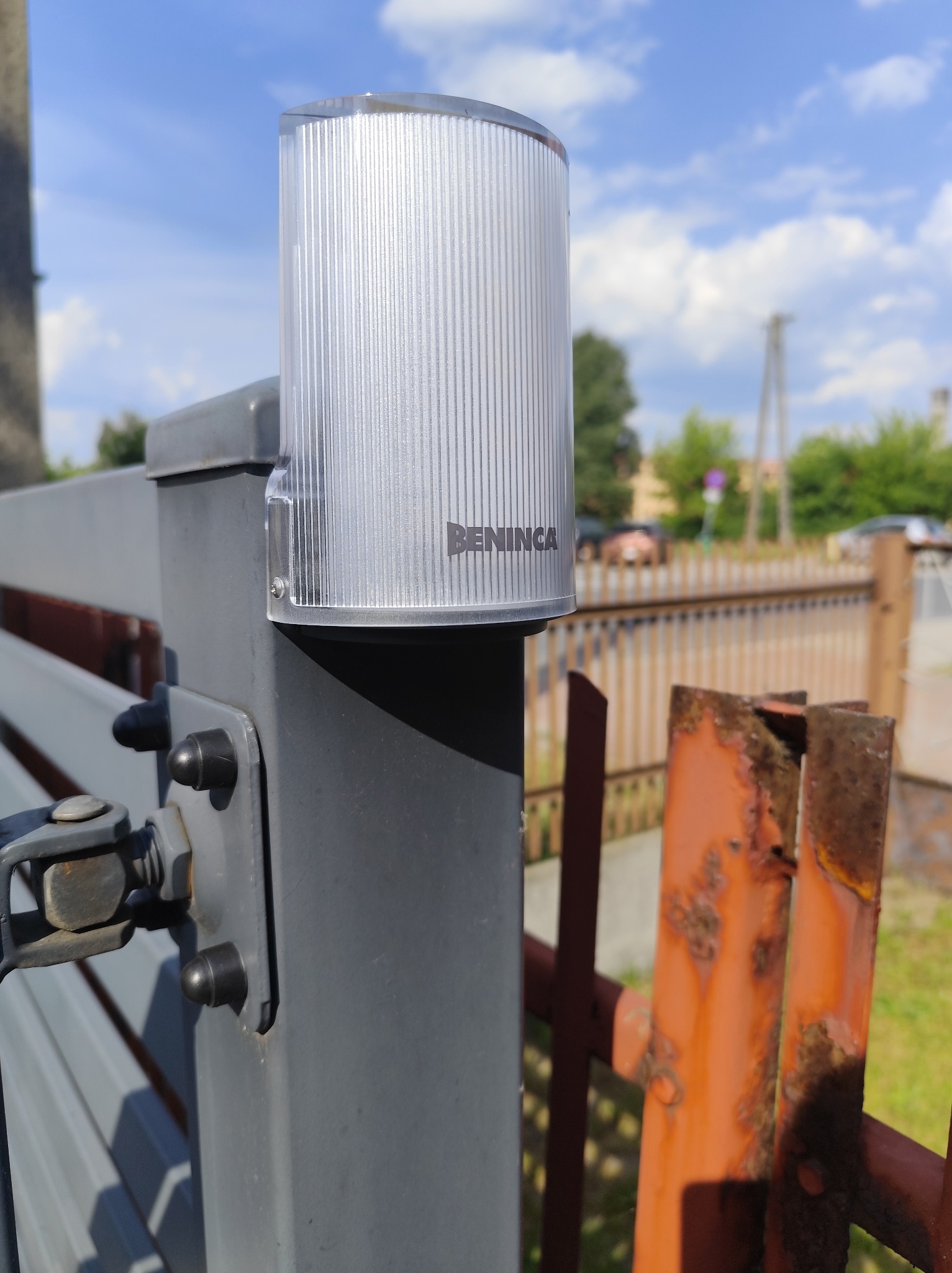 Lampa ostrzegawcza Beninca zamontowana na szarym słupku bramy, widoczna z bliska na tle błękitnego nieba i zardzewiałego ogrodzenia.