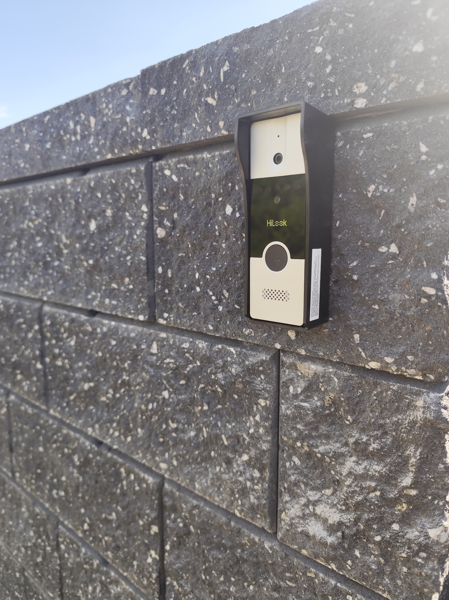 Zamontowany wideodomofon HiLook na szarej, teksturowanej ścianie z bloków. Kąt ujęcia z dołu podkreśla nowoczesny design urządzenia i solidność wykonania.