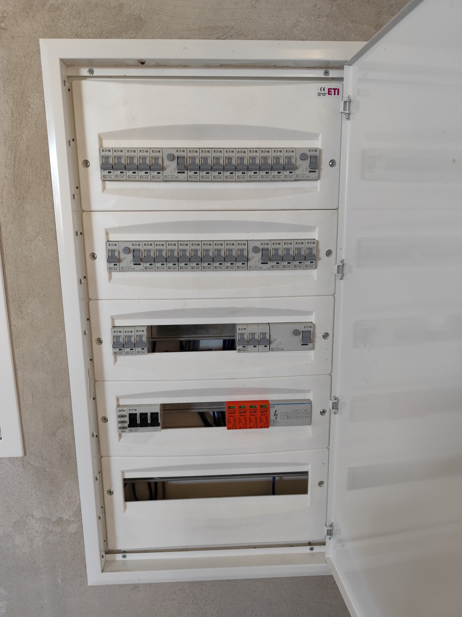Otwarta, biała skrzynka elektryczna z zainstalowanymi bezpiecznikami Eaton i ETI, widoczna na tle szarej ściany. Układ bezpieczników wewnątrz, widoczne przewody.