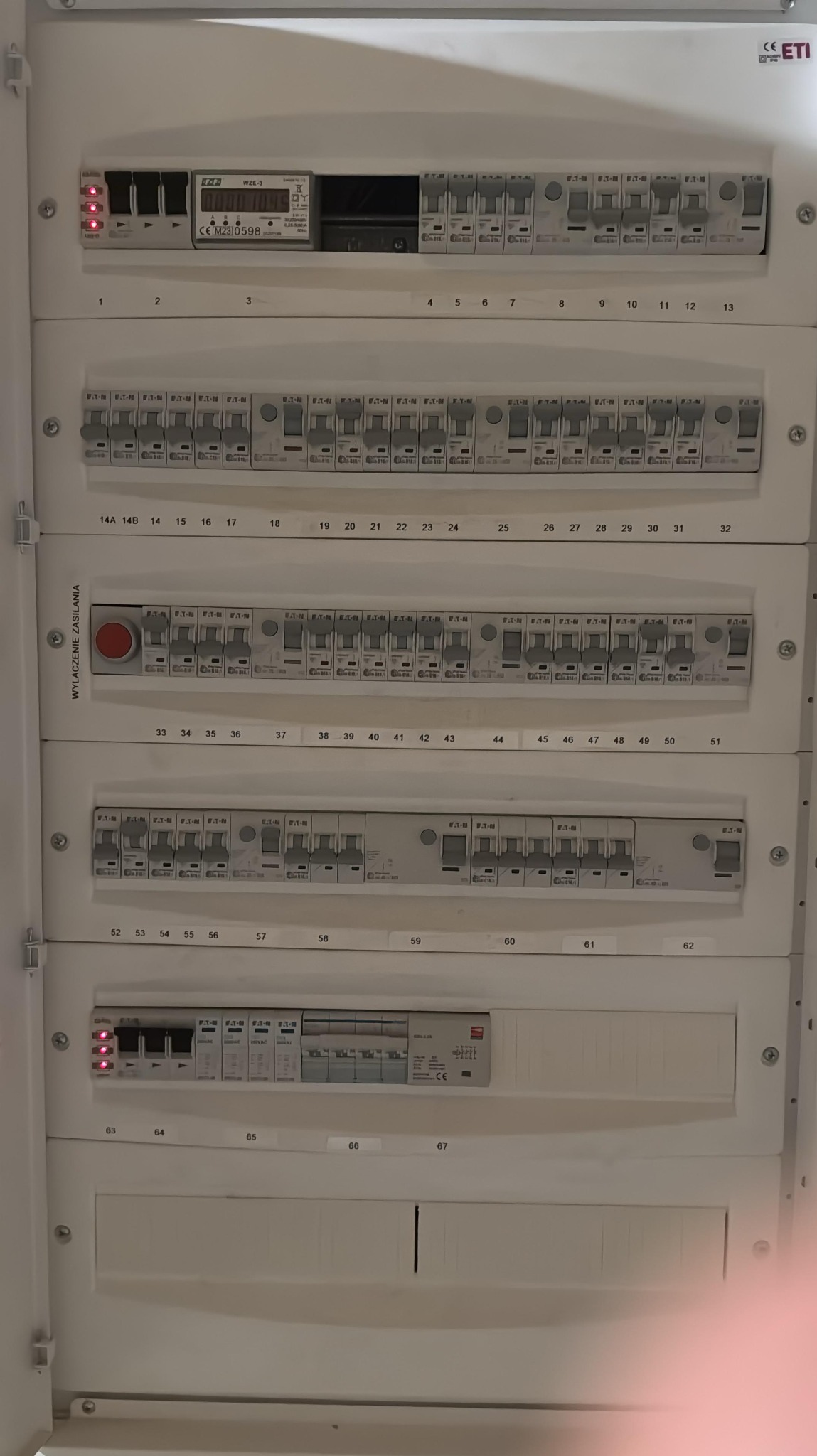 Rozdzielnica elektryczna z numerowanymi bezpiecznikami i licznikiem, widoczne diody kontrolne i oznaczenia producentów. Całość w białej obudowie.