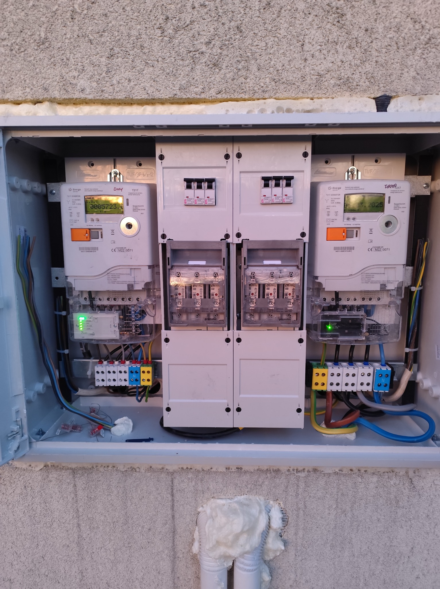 Otwarta skrzynka z dwoma licznikami energii elektrycznej i zabezpieczeniami, widoczne przewody, kostki elektryczne i elementy montażowe. Instalacja w szarej obudowie na szarej ścianie.