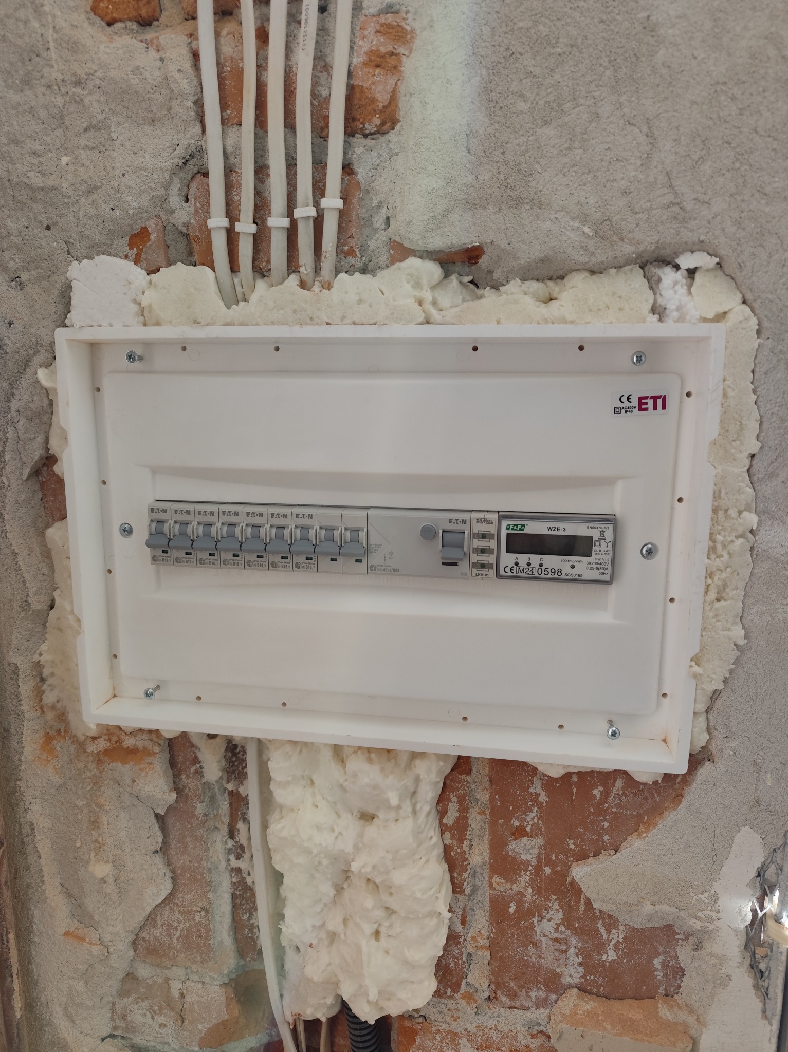 Zamontowana biała skrzynka elektryczna ETI z zabezpieczeniami, osadzona w surowej ścianie z cegły i betonu, uszczelniona pianką montażową. Widoczne kable w peszlach.
