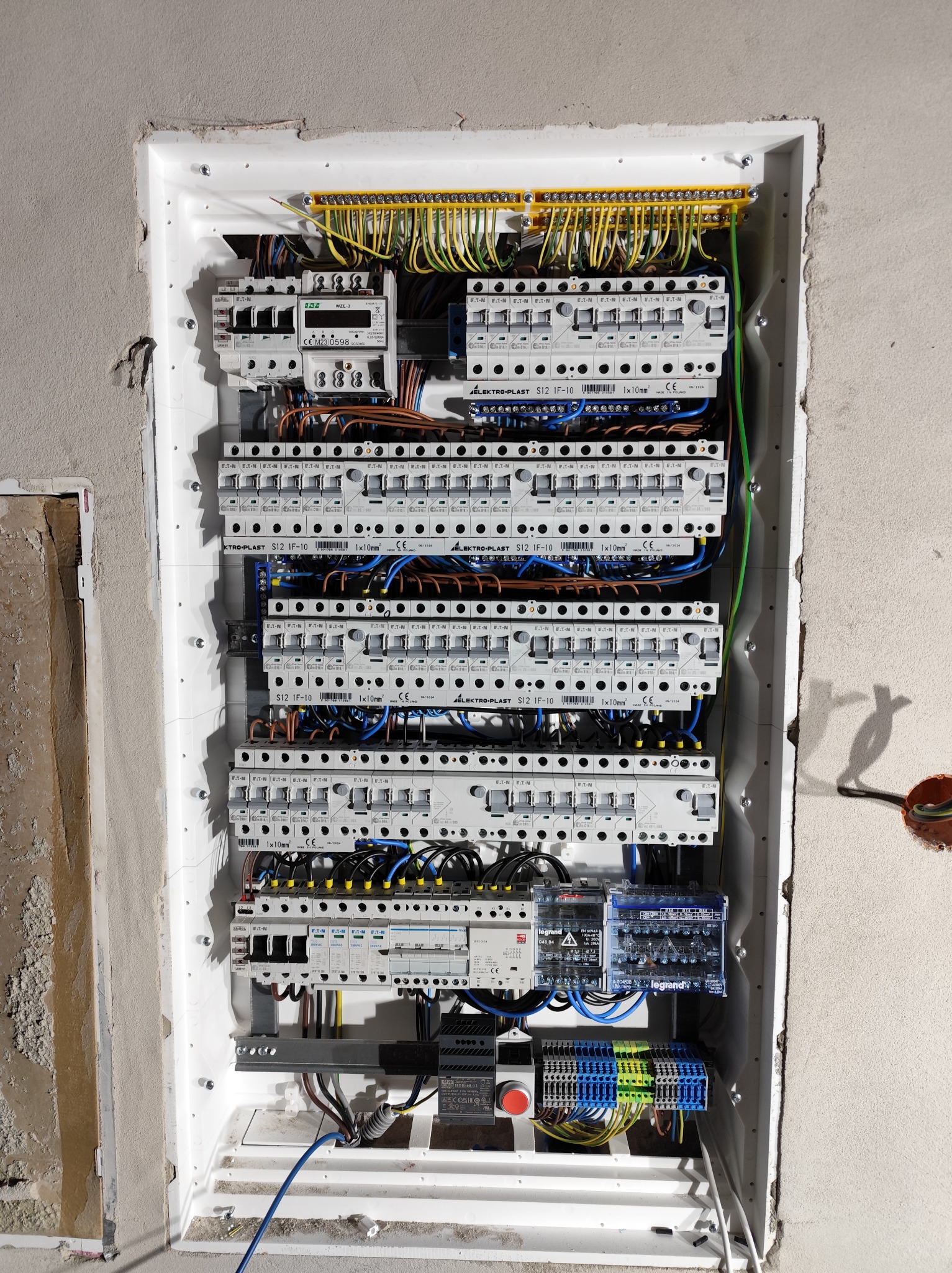 Rozdzielnica elektryczna w trakcie montażu, widoczne liczne zabezpieczenia, kable i złącza. Biała obudowa w surowej ścianie. Etap prac instalacyjnych.