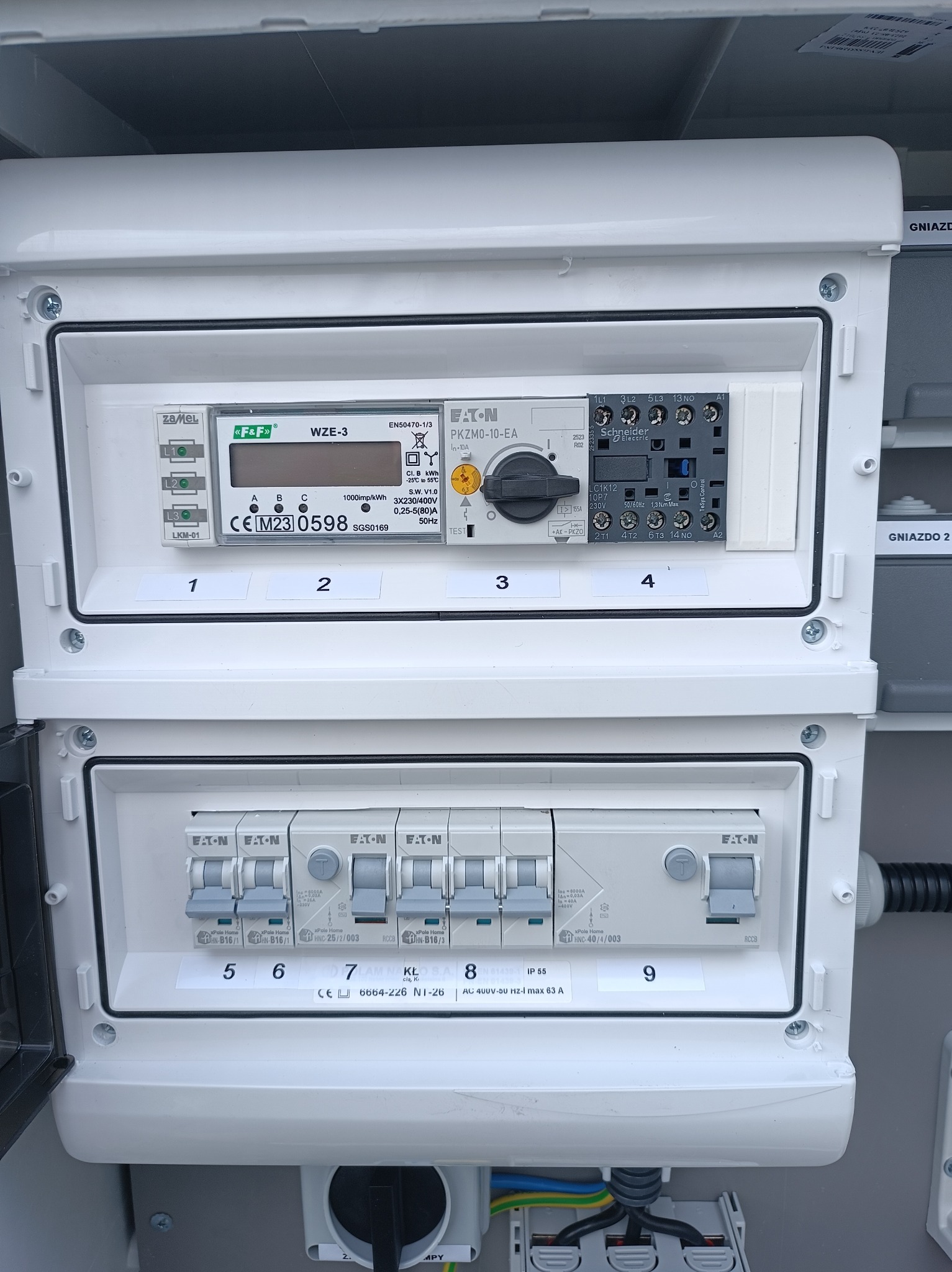 Rozdzielnia elektryczna z licznikiem i wyłącznikami firmy Eaton. Widoczne oznaczenia i parametry urządzeń. Skrzynka w kolorze białym, zamontowana wewnątrz budynku.