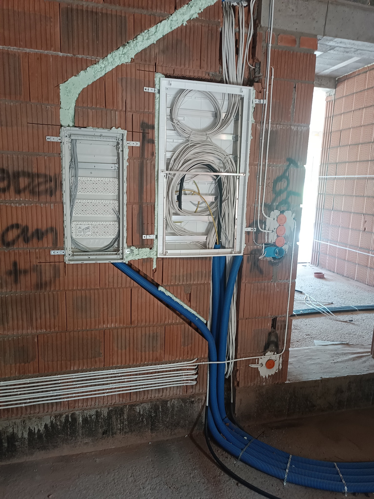 Rozdzielnie elektryczne z okablowaniem w surowym wnętrzu. Widoczne kable w peszlach i puszki instalacyjne na ścianie z czerwonej cegły. Instalacja w trakcie montażu.