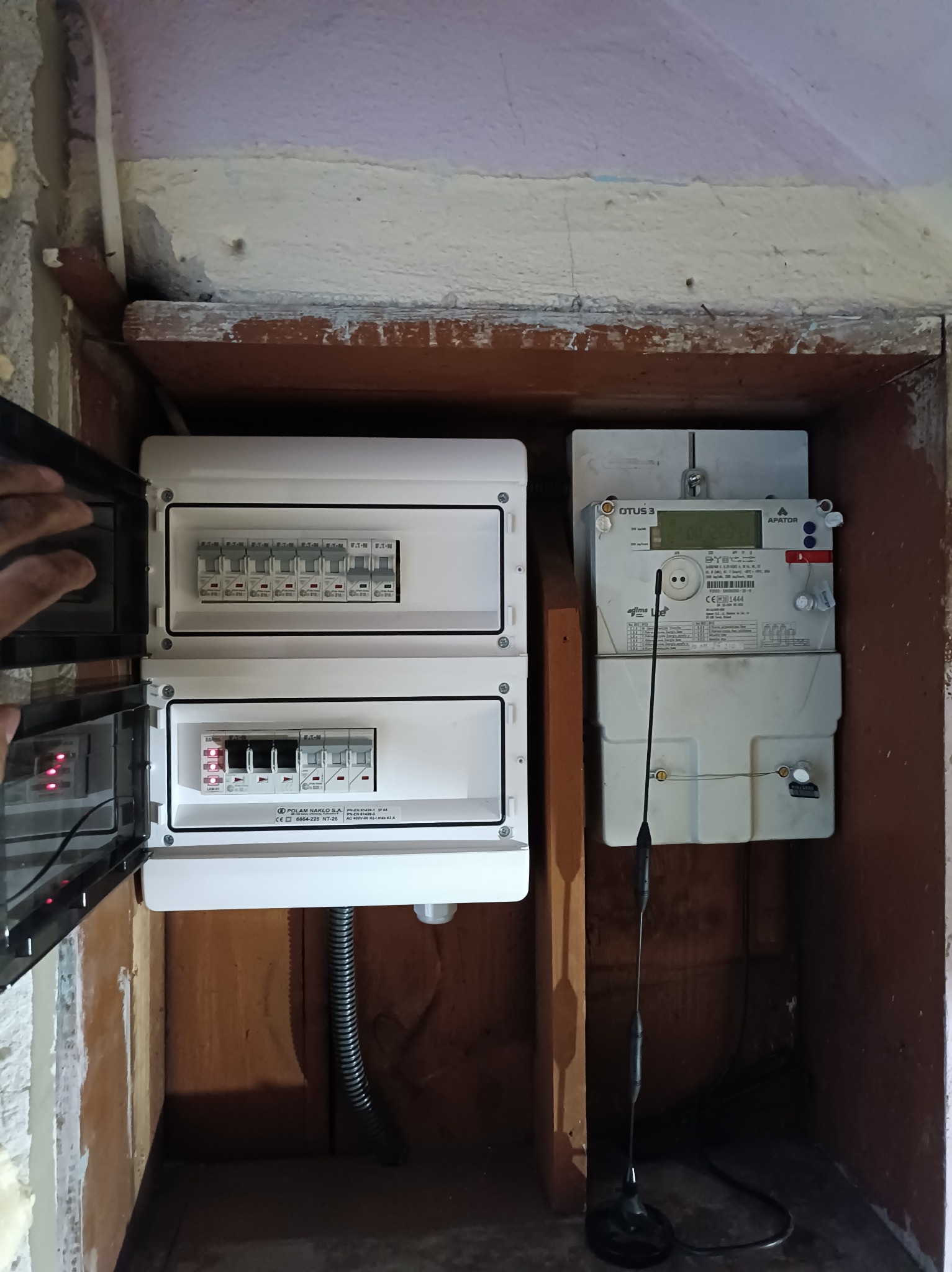 Zainstalowana skrzynka bezpiecznikowa z otwartymi drzwiczkami obok licznika energii elektrycznej w drewnianej wnęce. Widoczne zabezpieczenia i przewody.