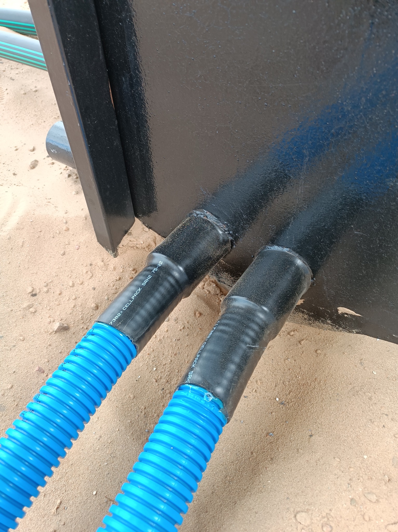 Dwa niebieskie, karbowane przewody wchodzą do czarnej skrzynki, zabezpieczone czarną taśmą termokurczliwą. Podłoże piaszczyste.