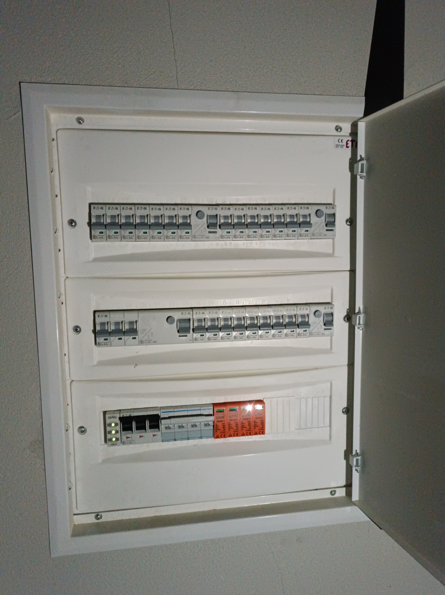 Otwarta, biała skrzynka elektryczna z bezpiecznikami EATON na szarej ścianie. Widoczne trzy rzędy bezpieczników i pomarańczowa tabliczka ostrzegawcza. Drzwiczki lekko uchylone.