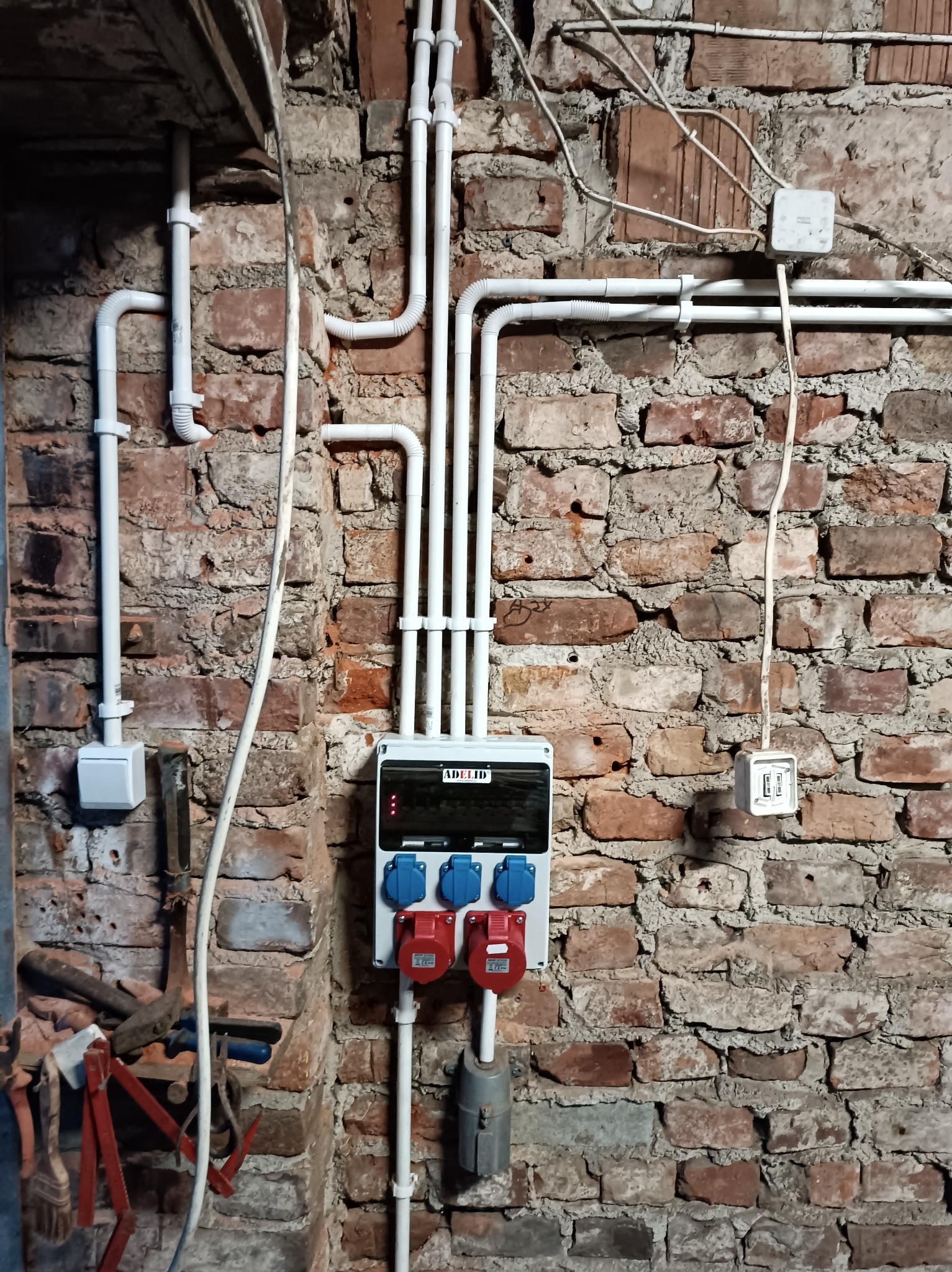 Instalacja elektryczna: białe rury PCV prowadzą kable wzdłuż ceglanej ściany do rozdzielnicy z gniazdami siłowymi. Widoczne puszki i narzędzia.