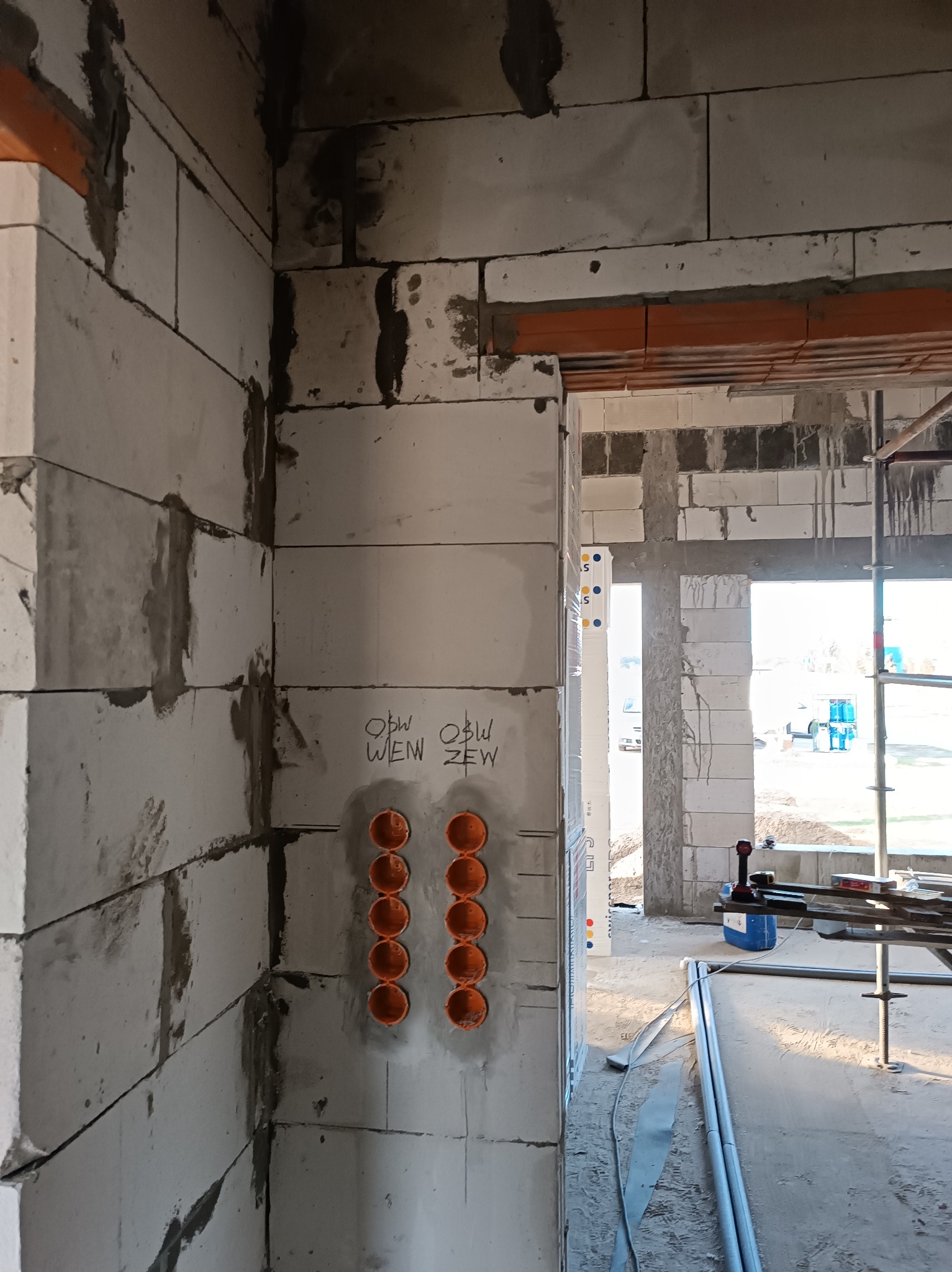 Wewnętrzny róg surowej ściany z betonu komórkowego z pomarańczowymi puszkami elektrycznymi podtynkowymi. Widoczne napisy na ścianie i elementy konstrukcyjne w tle.