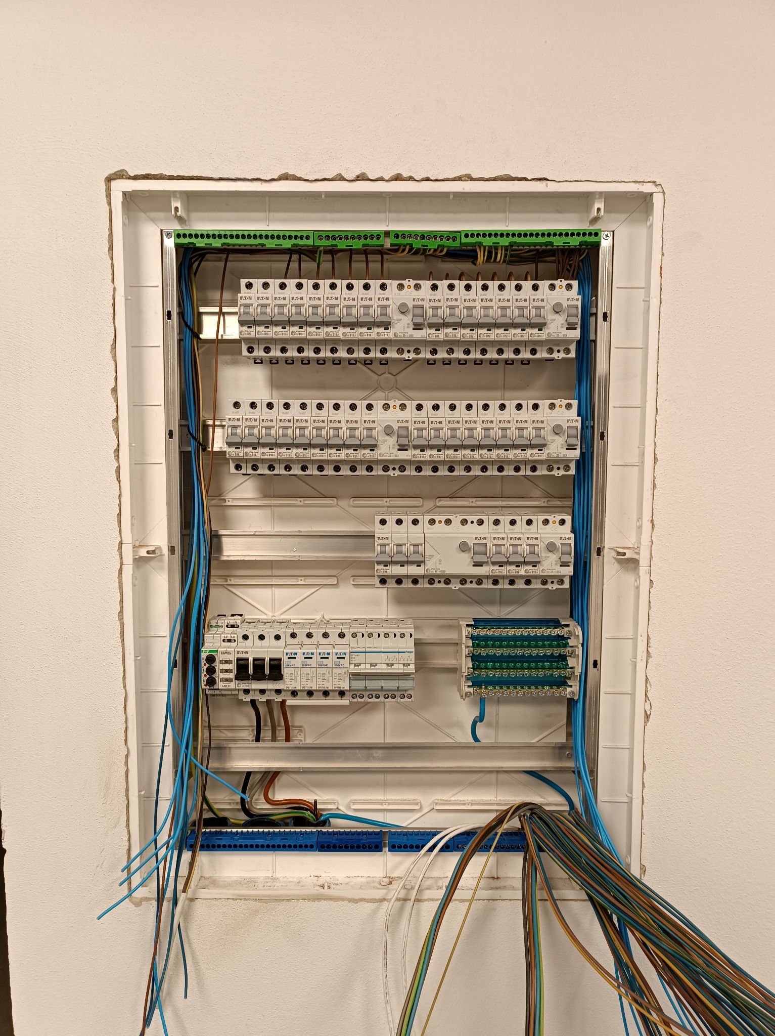 Rozdzielnica elektryczna w trakcie montażu, widoczne przewody i zabezpieczenia. Biała obudowa w surowej ścianie. Praca instalatora elektrycznego.