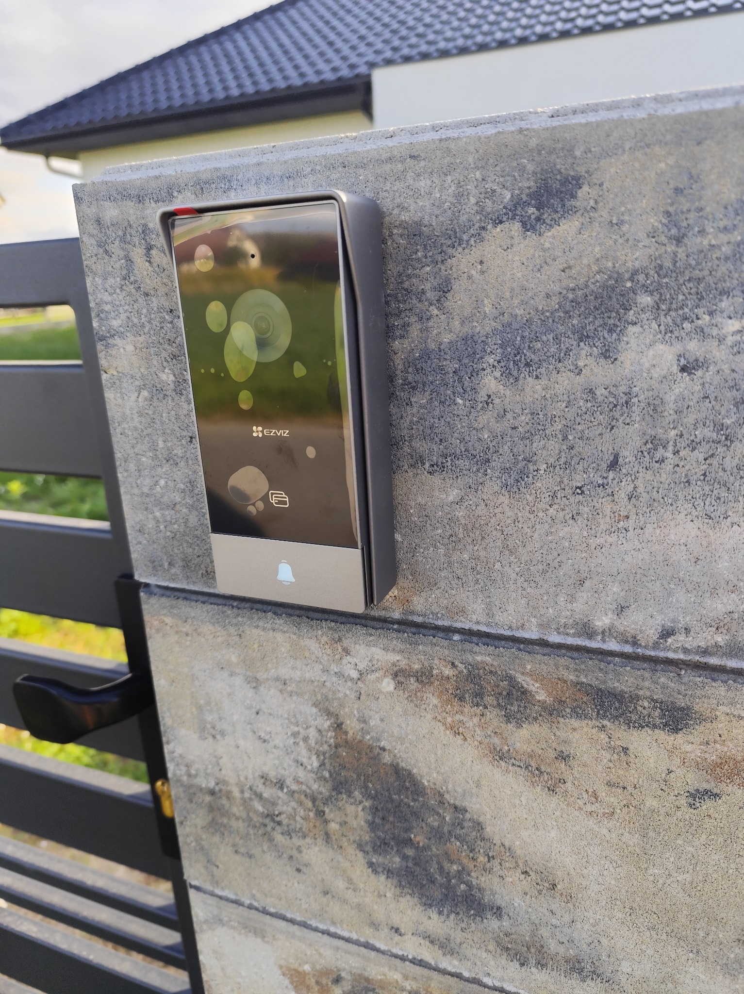 Nowoczesny wideodomofon EZVIZ zamontowany na kamiennym słupku bramy wjazdowej, widoczny z bliska. Słup z szaroburego kamienia, brama metalowa, w tle dom.