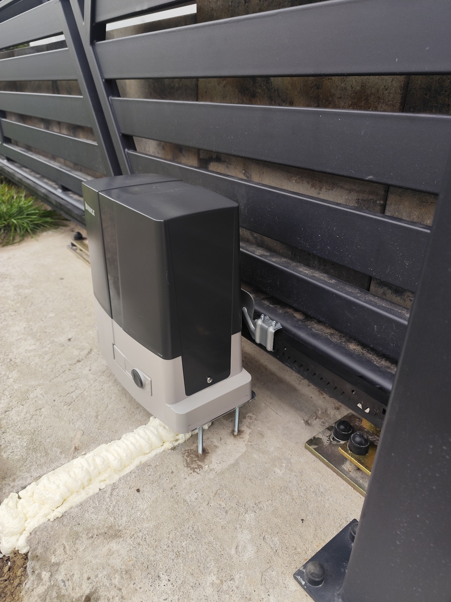 Zamontowany automat do bramy przesuwnej, widoczne mocowania i pianka montażowa. Brama w kolorze grafitowym.