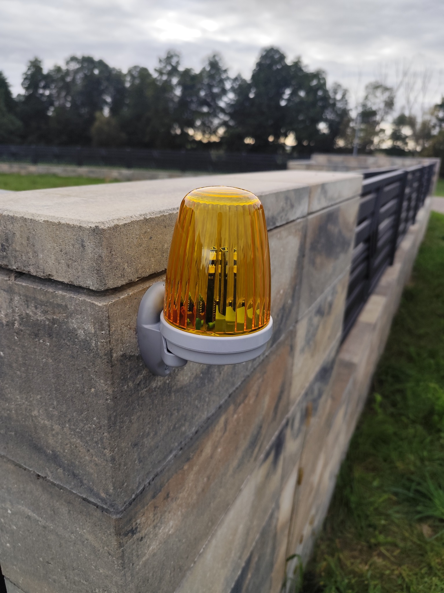 Zamontowana lampa ostrzegawcza, z żółtym kloszem, na betonowym słupku ogrodzenia z metalowymi przęsłami. Tło stanowią drzewa i pochmurne niebo.