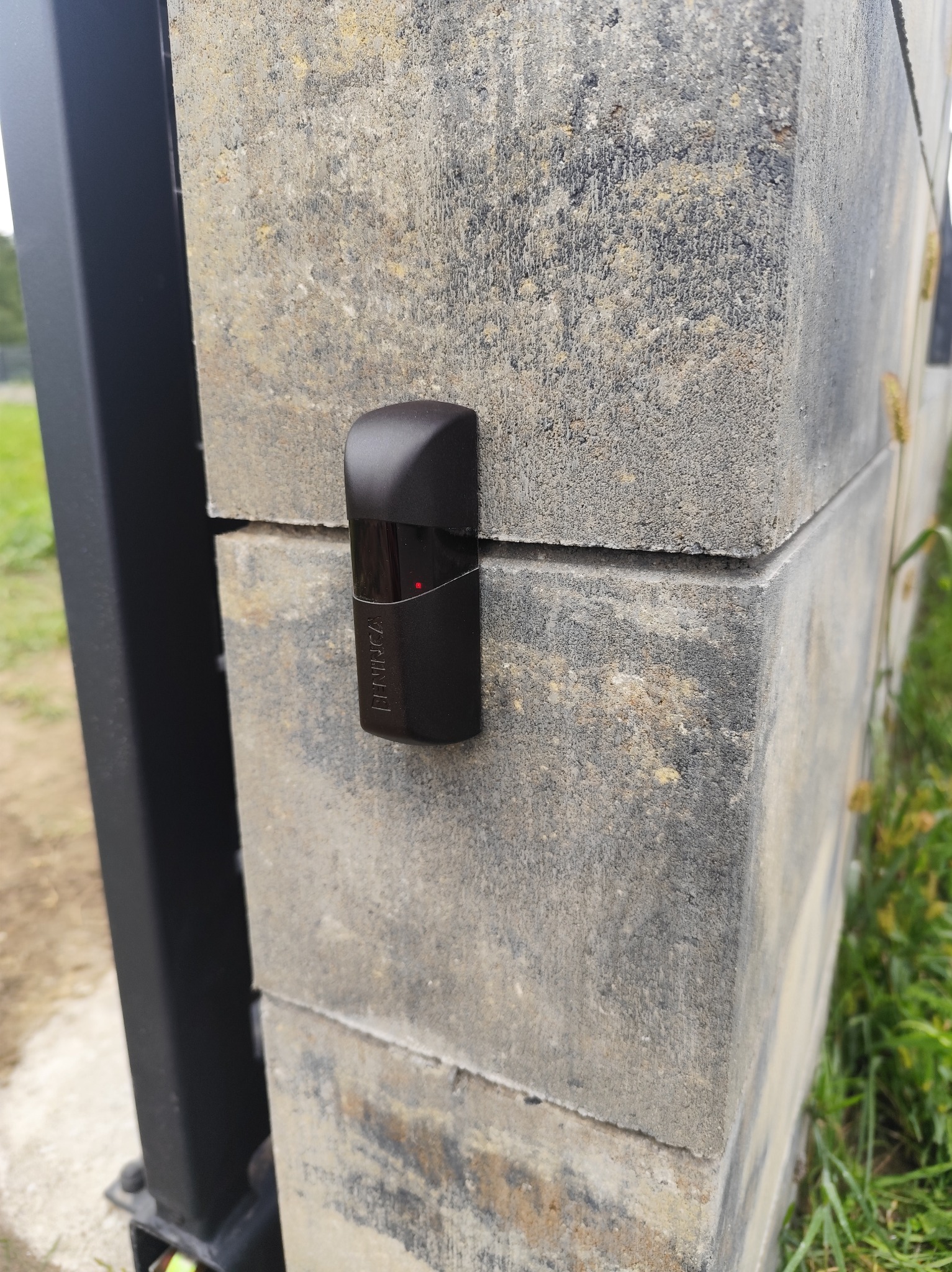 Czujnik zbliżeniowy marki Beninca zamontowany na słupku bramy z betonu architektonicznego. Widoczny element konstrukcji bramy i fragment zieleni w tle.