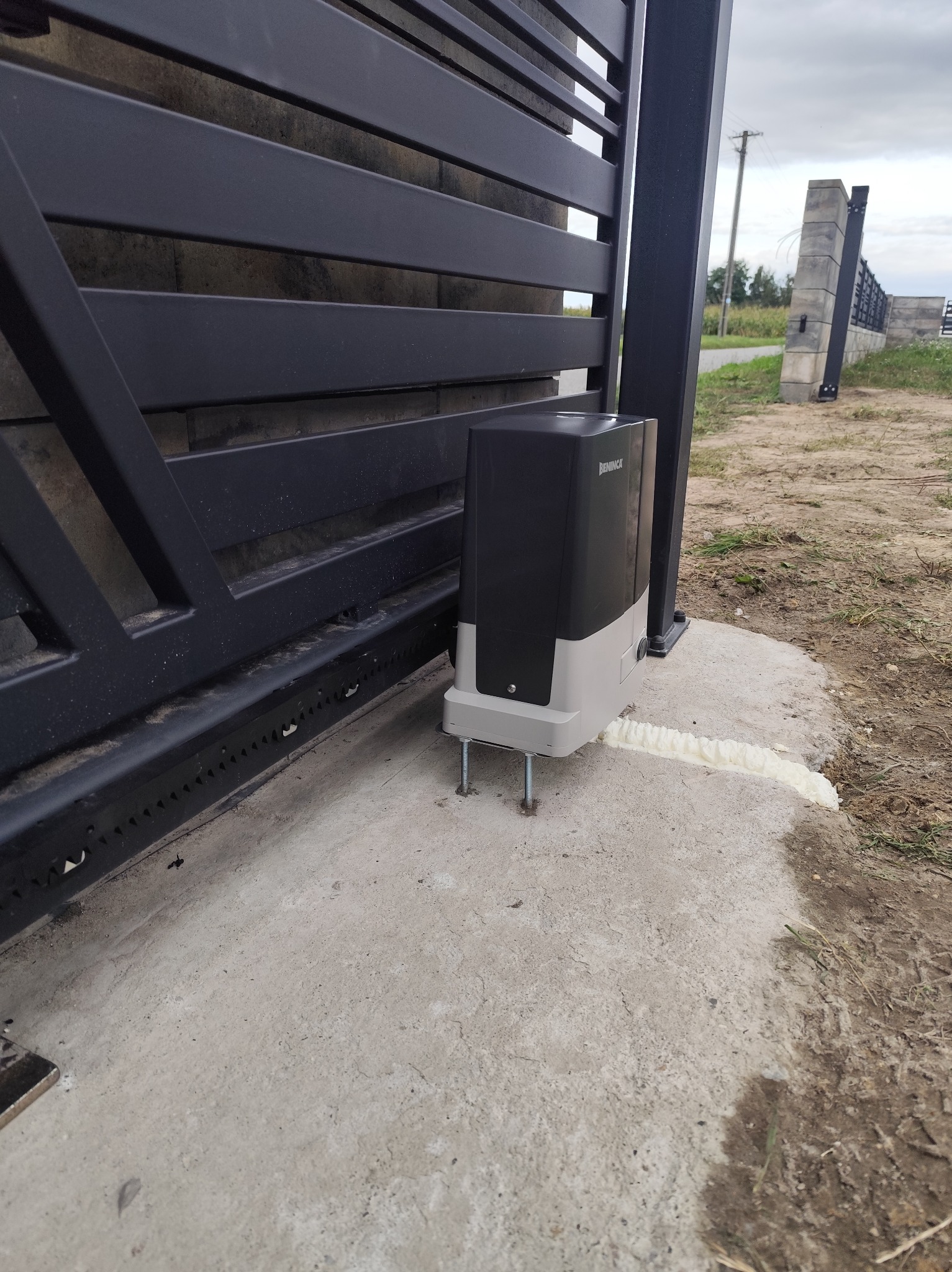 Zamontowany napęd do bramy przesuwnej marki Beninca, widoczny na betonowej podstawie, z pianką montażową uszczelniającą szczelinę. W tle fragment bramy i słupki.