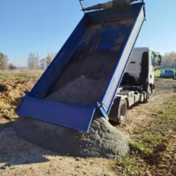 Ciężarówka z podniesioną skrzynią ładunkową wysypuje żwir na placu budowy w słoneczny dzień.
