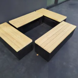 Cztery modułowe ławki z jasnego drewna na czarnych podstawach, ustawione w kwadrat, na szarym podłożu, z widocznym fragmentem ściany i okna w tle.