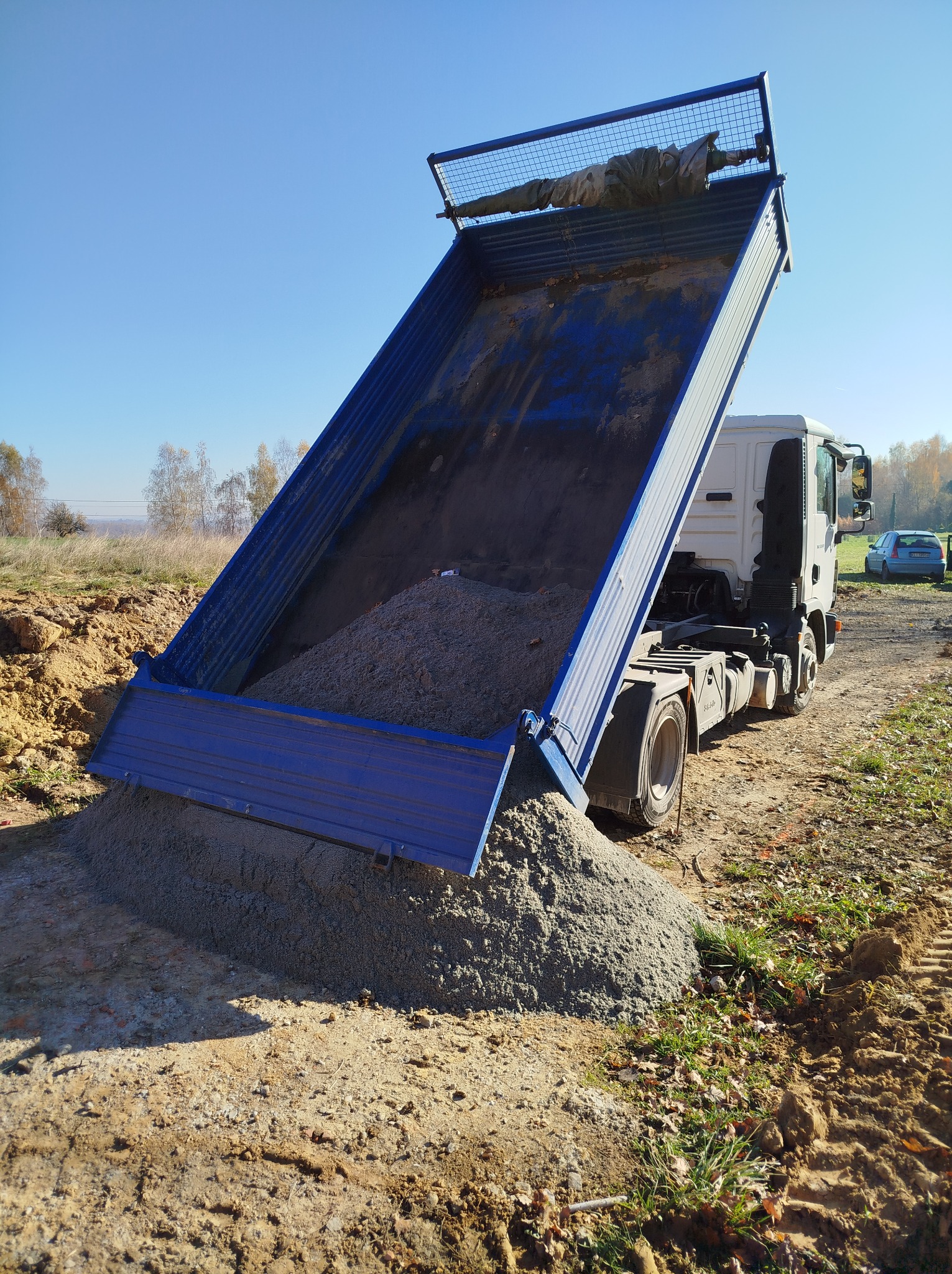 Ciężarówka z podniesioną skrzynią ładunkową wysypuje żwir na placu budowy w słoneczny dzień.