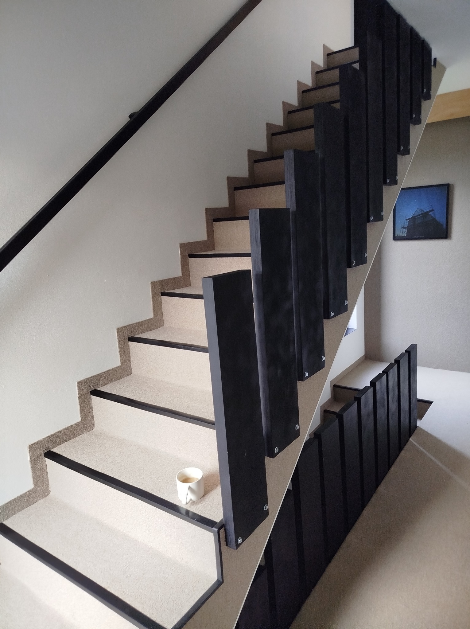 Nowoczesne schody wewnętrzne z jasną wykładziną i czarnymi elementami wykończeniowymi, na jednym ze stopni stoi biały kubek z kawą.
