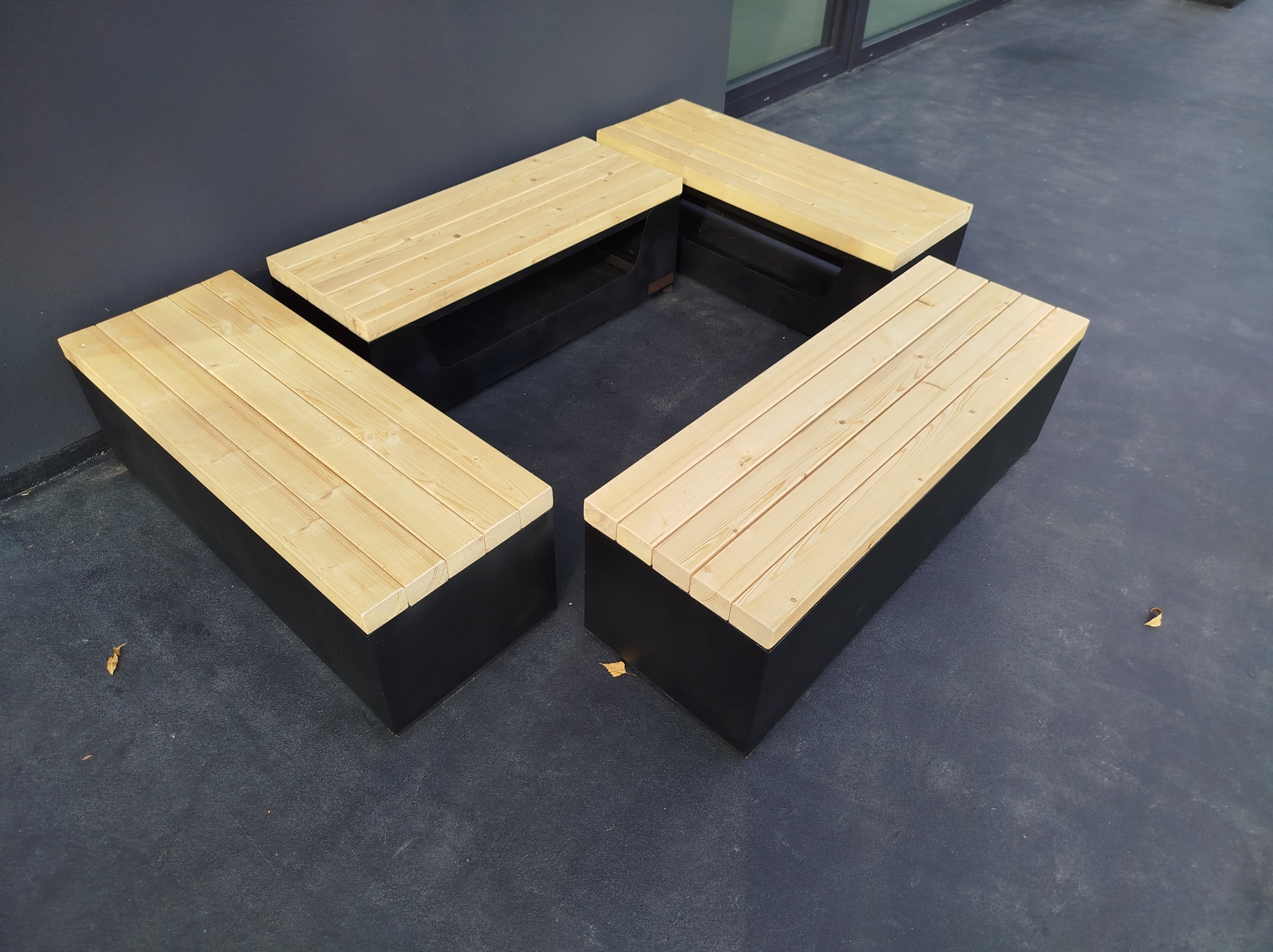 Cztery modułowe ławki z jasnego drewna na czarnych podstawach, ustawione w kwadrat, na szarym podłożu, z widocznym fragmentem ściany i okna w tle.