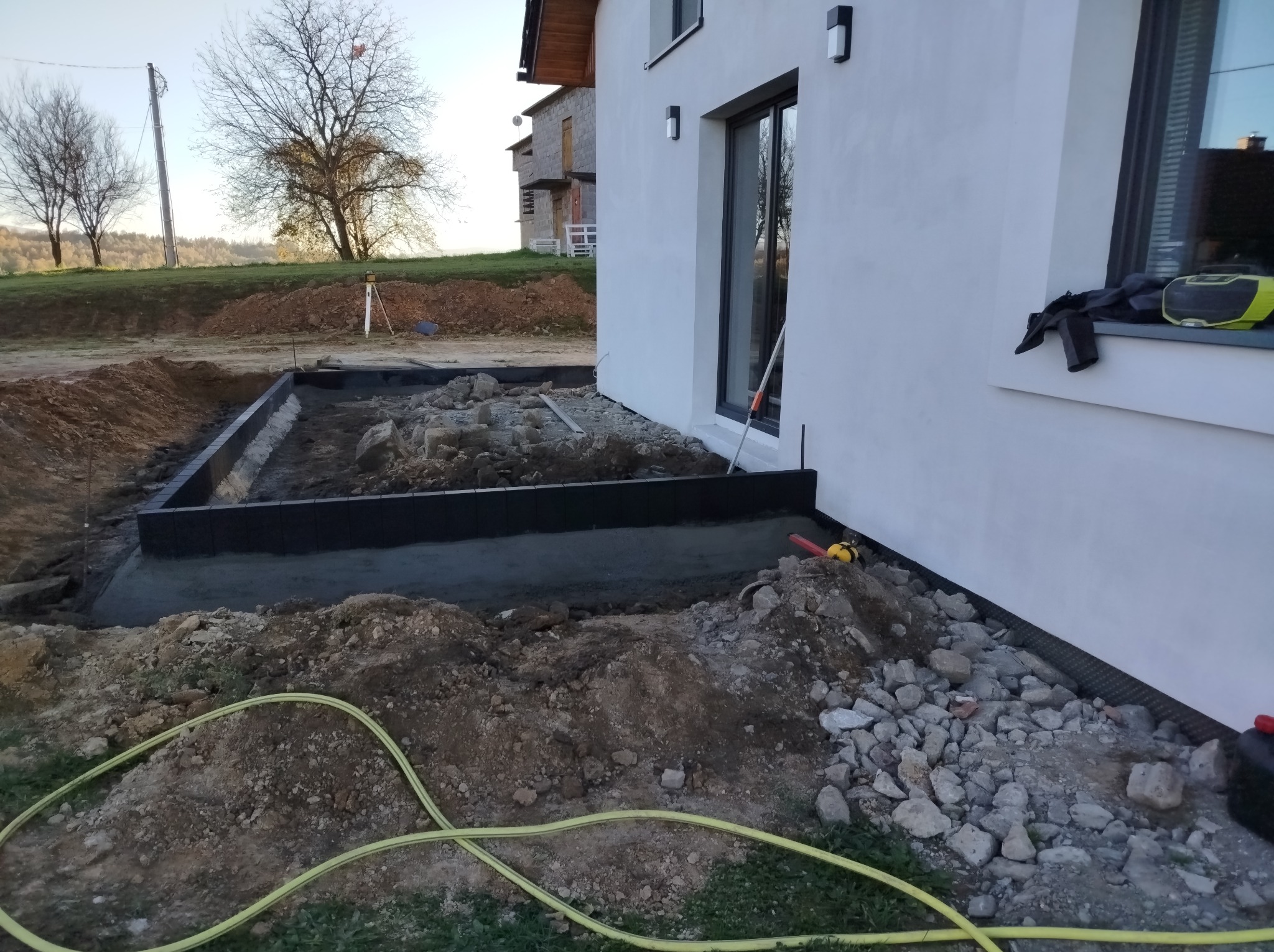 Przygotowany wykop pod fundamenty budynku z czarną izolacją pionową, widoczny żółty wąż ogrodowy i niwelator optyczny w tle, teren budowy w trakcie prac.
