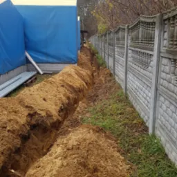 Długi, wąski wykop w ziemi, biegnący wzdłuż betonowego ogrodzenia z dekoracyjnymi elementami, obok tymczasowej konstrukcji z niebieskiej plandeki i rur