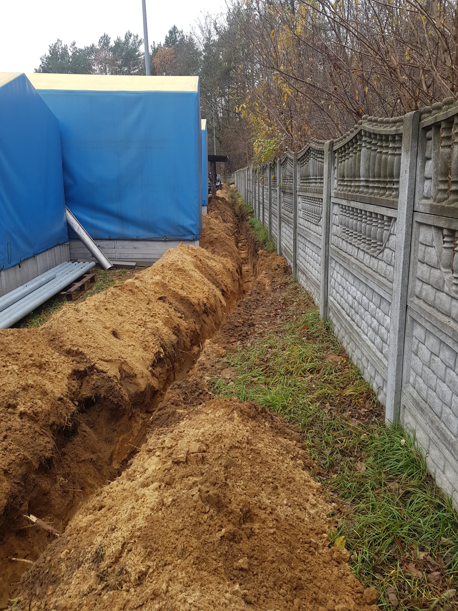 Długi, wąski wykop w ziemi, biegnący wzdłuż betonowego ogrodzenia z dekoracyjnymi elementami, obok tymczasowej konstrukcji z niebieskiej plandeki i rur