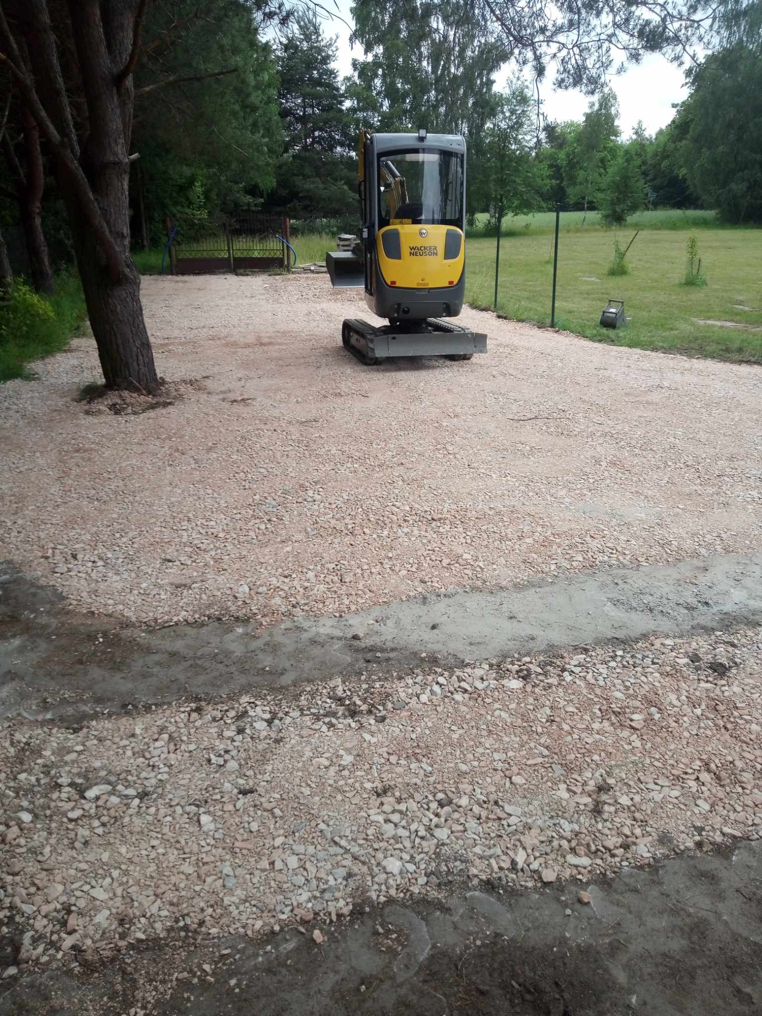 Żółta mini koparka Wacker Neuson na wyrównanym terenie z jasnego kruszywa, widoczne ślady opon i przygotowane podłoże pod kostkę, w tle zieleń drzew i ogrodzenie.
