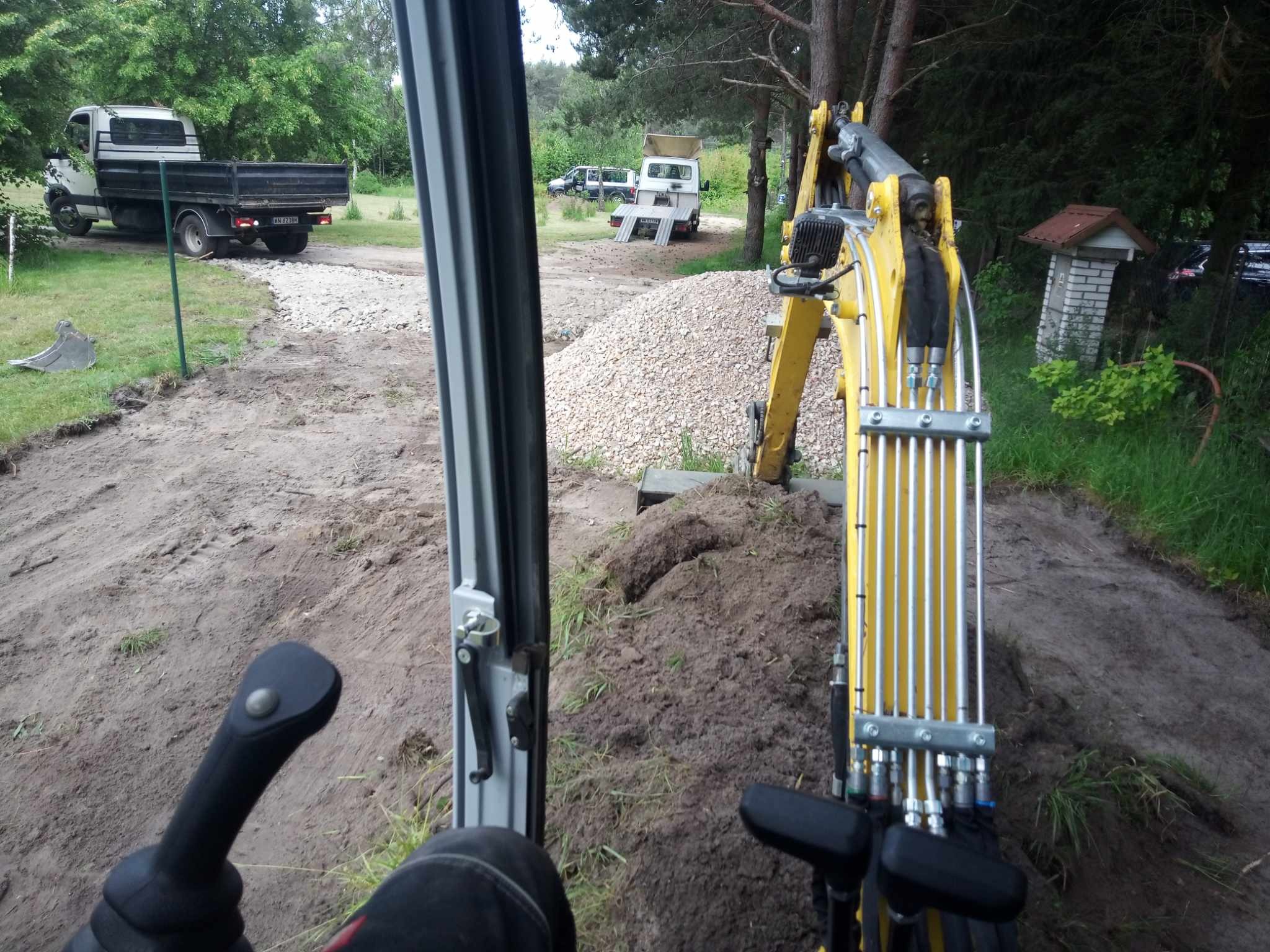 Widok z kabiny żółtej minikoparki w Dobre. W tle hałda żwiru i zaparkowane samochody dostawcze. Widoczne elementy sterujące koparki.