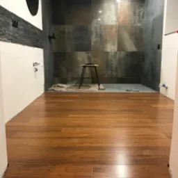 Wnętrze łazienki w trakcie remontu: ułożona drewniana podłoga, ściany wyłożone wielkoformatowymi płytkami w odcieniach szarości i brązu, widoczne ślady prac wykończeniowych przy ścianach.