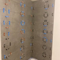 Hilton-Bud - Świeżo ułożone, duże, szare płytki w rogu kabiny prysznicowej, z kolorowymi krzyżykami dystansowymi: niebieskimi, brązowymi i czarnymi, utrzymującymi równe odstępy.