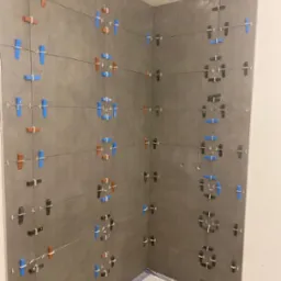 Świeżo ułożone, duże, szare płytki w rogu kabiny prysznicowej, z kolorowymi krzyżykami dystansowymi: niebieskimi, brązowymi i czarnymi, utrzymującymi równe odstępy.