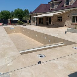 Hilton-Bud - Wyłożony beżowymi płytkami basen w trakcie budowy, z widocznymi otworami na oświetlenie i odpływy. W tle dom z cegły i zielenią. Słoneczny dzień.