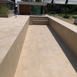 Hilton-Bud - Wyłożony beżowymi płytkami basen w ogrodzie, z widocznymi schodkami i oświetleniem w ścianach. W tle budynek z drewnianą elewacją i zielenią.