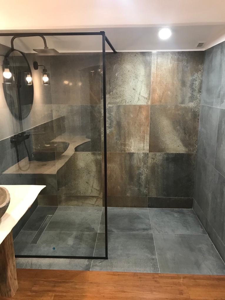 Nowoczesna kabina prysznicowa z ciemnymi płytkami imitującymi beton, czarną armaturą i okrągłym lustrem z oświetleniem punktowym, uzupełniona drewnianą podłogą i blatem.