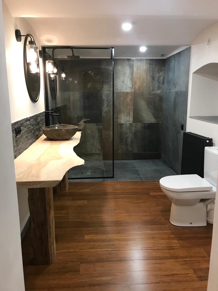 Nowoczesna łazienka z drewnianą podłogą, umywalką z kamienia na blacie z surowego drewna, kabiną prysznicową z czarnymi ramami i płytkami imitującymi kamień.