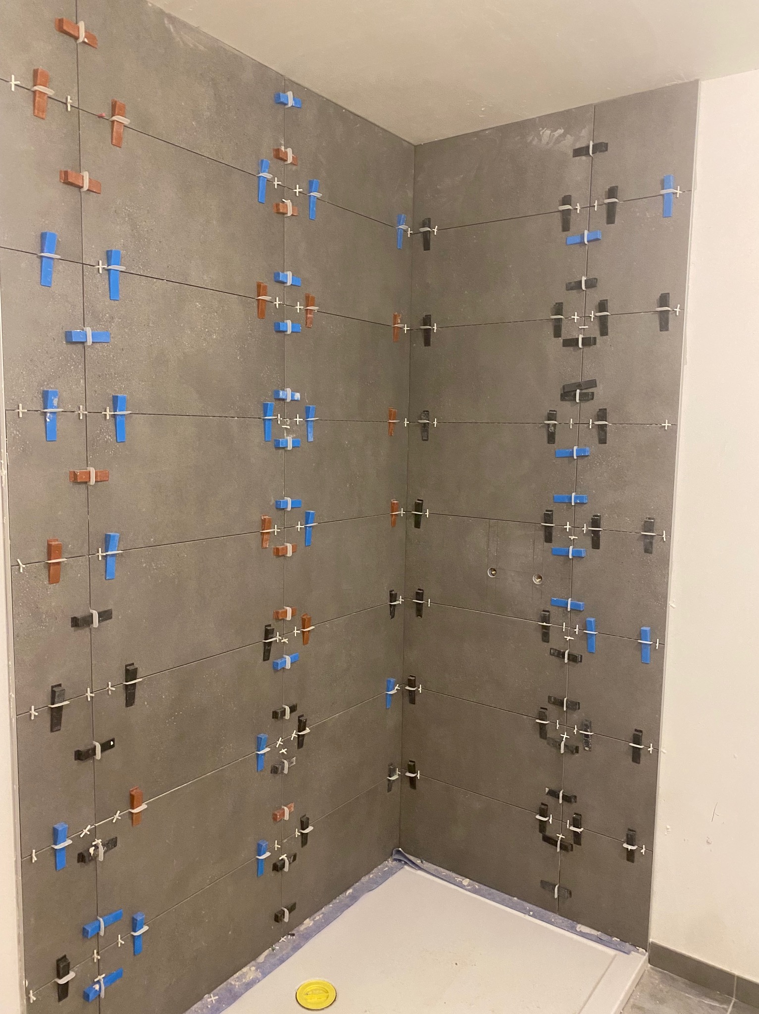 Świeżo ułożone, duże, szare płytki w rogu kabiny prysznicowej, z kolorowymi krzyżykami dystansowymi: niebieskimi, brązowymi i czarnymi, utrzymującymi równe odstępy.