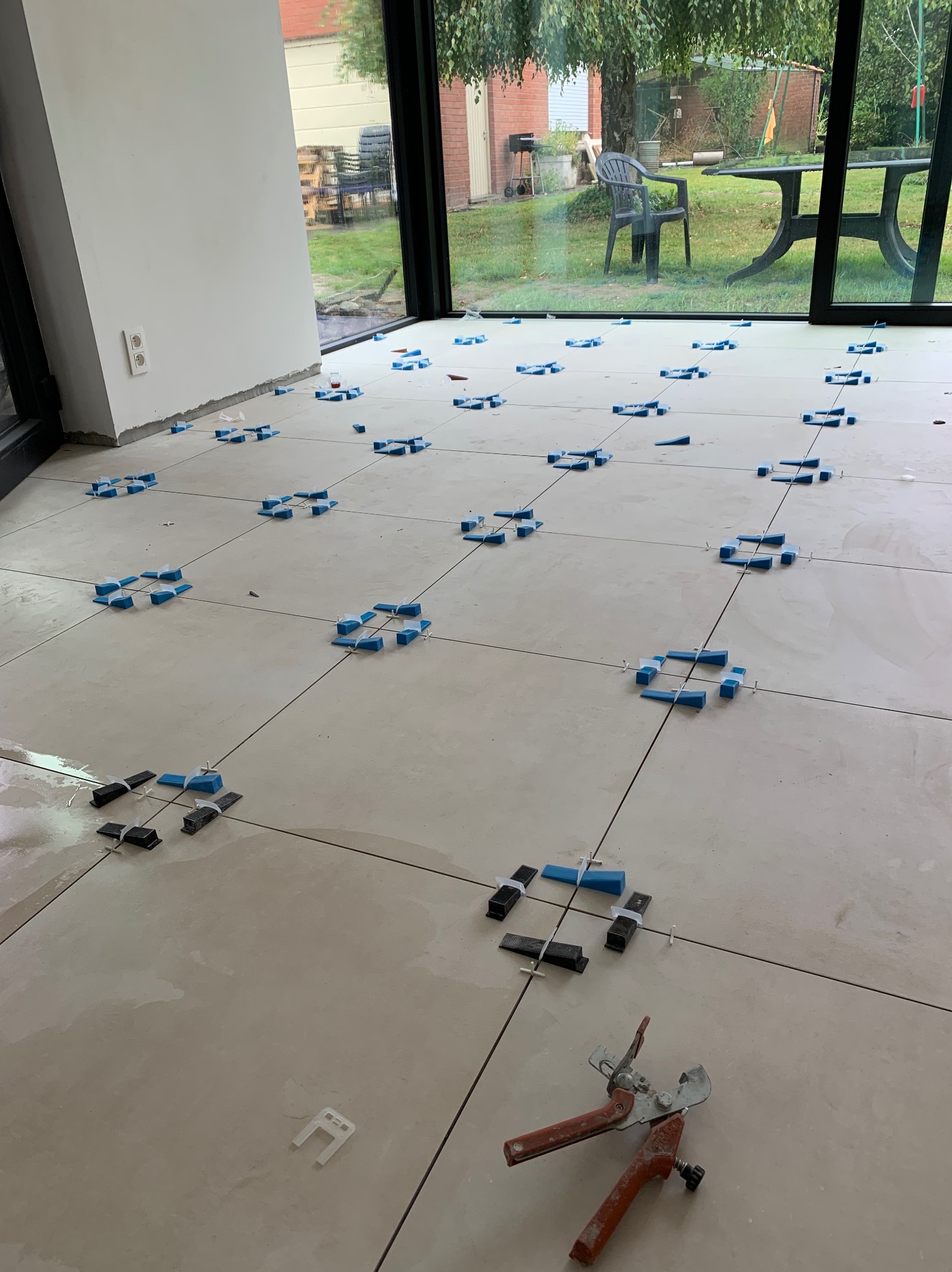 Układanie płytek podłogowych z użyciem systemu poziomowania, widoczne narzędzie do zaciskania klinów, duże okno z widokiem na ogród w tle.