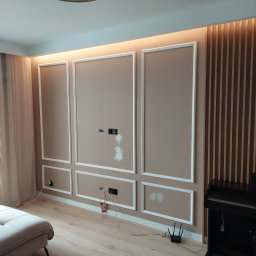 Piotr ciesiółka - Ściana w salonie z zamontowanymi panelami ściennymi w kolorze beżowym z białymi listwami dekoracyjnymi, podświetleniem LED i drewnianymi lamelami, widoczne gniazdka elektryczne i otwory po...